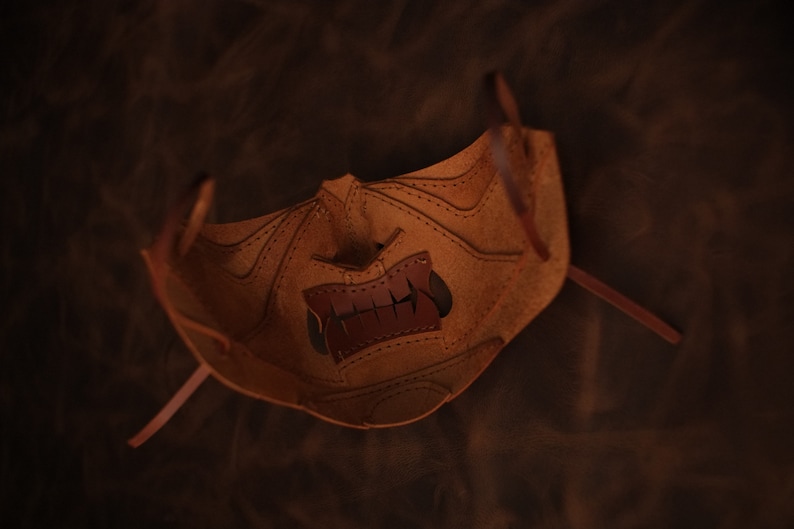 Pu&ograve; includere: Una maschera in pelle marrone con un design cucito e un'apertura per la bocca. La maschera ha due lunghi lacci in pelle per legare.