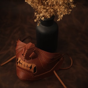 Pu&ograve; includere: Una maschera in pelle marrone con un design dettagliato, che presenta un ghigno dentato e cuciture intricate. La maschera &egrave; appoggiata su una superficie marrone, con un vaso di fiori secchi sullo sfondo.