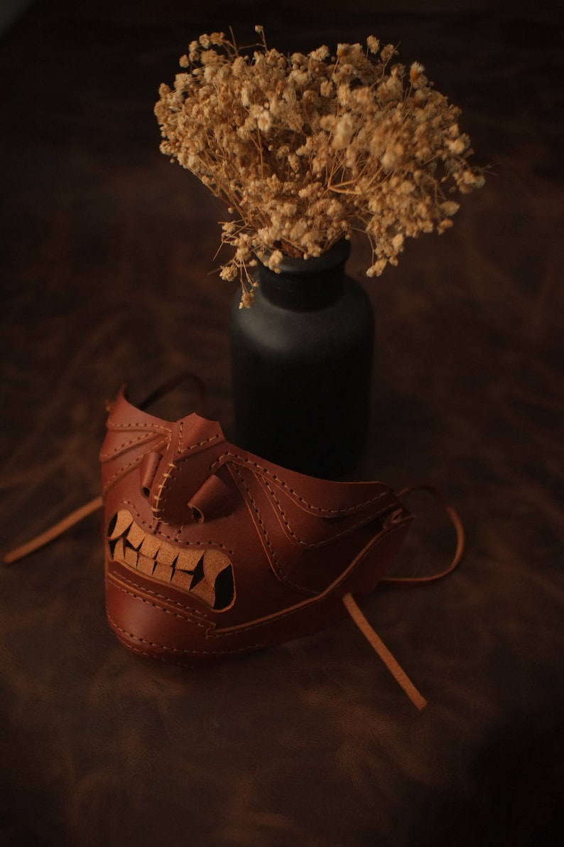 Pu&ograve; includere: Una maschera in pelle marrone con un design dettagliato, che presenta un ghigno dentato e cuciture intricate. La maschera &egrave; appoggiata su una superficie marrone, con un vaso di fiori secchi sullo sfondo.