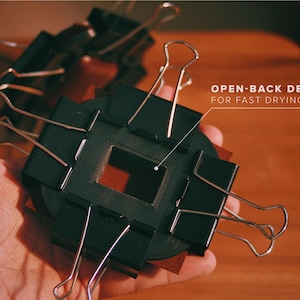 May include: A black open-back frame with a square opening, held in place by silver binder clips. The frame is resting on a brown surface. The text "OPEN-BACK DESIGN FOR FAST DRYING TIME" is visible in the upper right corner of the image.