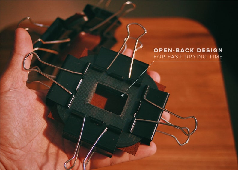 May include: A black open-back frame with a square opening, held in place by silver binder clips. The frame is resting on a brown surface. The text "OPEN-BACK DESIGN FOR FAST DRYING TIME" is visible in the upper right corner of the image.