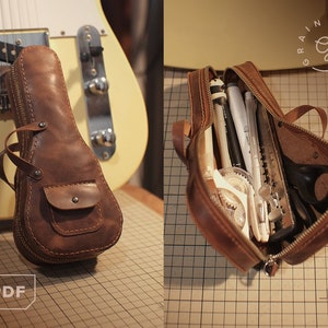 以下が含まれることがあります： ジッパー留めと前面に小さなポケットが付いた茶色の革製のウクレレケース。ケースは開いており、ペン、鉛筆、ハサミ、定規など、さまざまなクラフト用品が入っています。ケースはグリッド状の表面の上に置かれています。