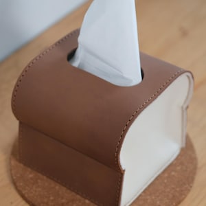 Pode incluir: Uma caixa de len&ccedil;os de papel castanha e branca com a parte superior arredondada e uma base retangular. A caixa &eacute; feita de um material semelhante a couro e tem um design costurado. Os len&ccedil;os de papel brancos s&atilde;o vis&iacute;veis, emergindo da abertura superior. A caixa est&aacute; sobre um porta-copos de corti&ccedil;a.
