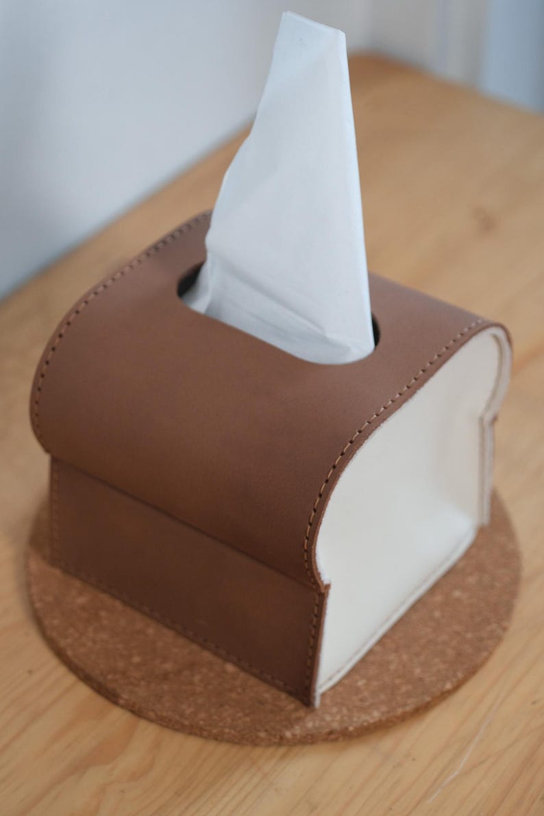 Pode incluir: Uma caixa de len&ccedil;os de papel castanha e branca com a parte superior arredondada e uma base retangular. A caixa &eacute; feita de um material semelhante a couro e tem um design costurado. Os len&ccedil;os de papel brancos s&atilde;o vis&iacute;veis, emergindo da abertura superior. A caixa est&aacute; sobre um porta-copos de corti&ccedil;a.