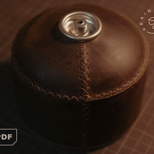 May include: A brown leather canister with a stitched seam around the top and a silver metal cap. The canister is sitting on a brown surface.