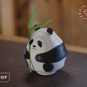 Peut inclure: Figurine de panda artisanal en cuir blanc et noir, tenant une tige de bambou verte. Le panda a des yeux, des oreilles et une tache ventrale noirs. La figurine est sur une surface marron.