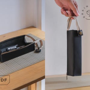May include: Black leather pencil case with a zip and a beige handle. The rectangular case is open, showing a pen and pencil inside. The case is displayed on a wooden surface.