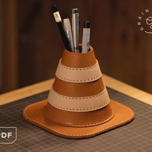 Könnte beinhalten: Ein handgefertigter Schreibtisch-Organizer aus Leder in Kegelform mit quadratischer Basis. Der Organizer besteht aus braunem und beigem Leder mit einem genähten Design. Mehrere Stifte und Bleistifte befinden sich im Inneren. Das PDF-Logo befindet sich unten links.