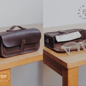 May include: A dark brown leather eyeglasses case with a handle and a snap closure. The case is shown closed and open, revealing a pair of clear-framed glasses and a cleaning cloth. The case is sitting on a wooden surface.