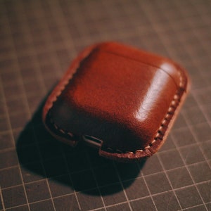 May include: A brown leather case for wireless earbuds. The case has a stitched seam around the edge and a small opening for charging.