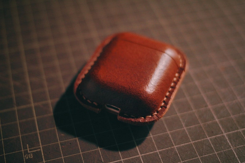 May include: A brown leather case for wireless earbuds. The case has a stitched seam around the edge and a small opening for charging.