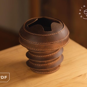 May include: A brown leather container with a screw-like design. The container has a circular top with a cutout and is made of stacked, stitched leather rings. The item is on a wooden surface.