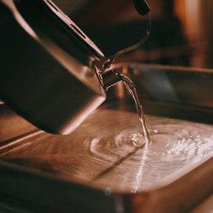 May include: A silver kettle pouring water into a stainless steel tray, creating a swirling pattern in the water.