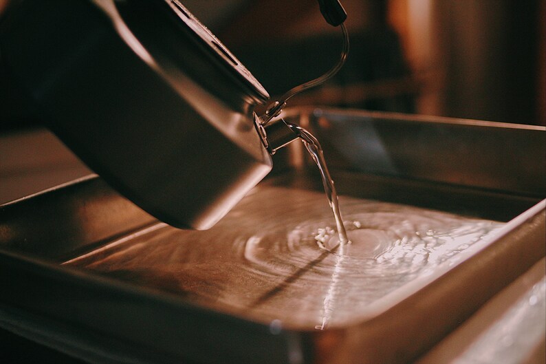 May include: A silver kettle pouring water into a stainless steel tray, creating a swirling pattern in the water.