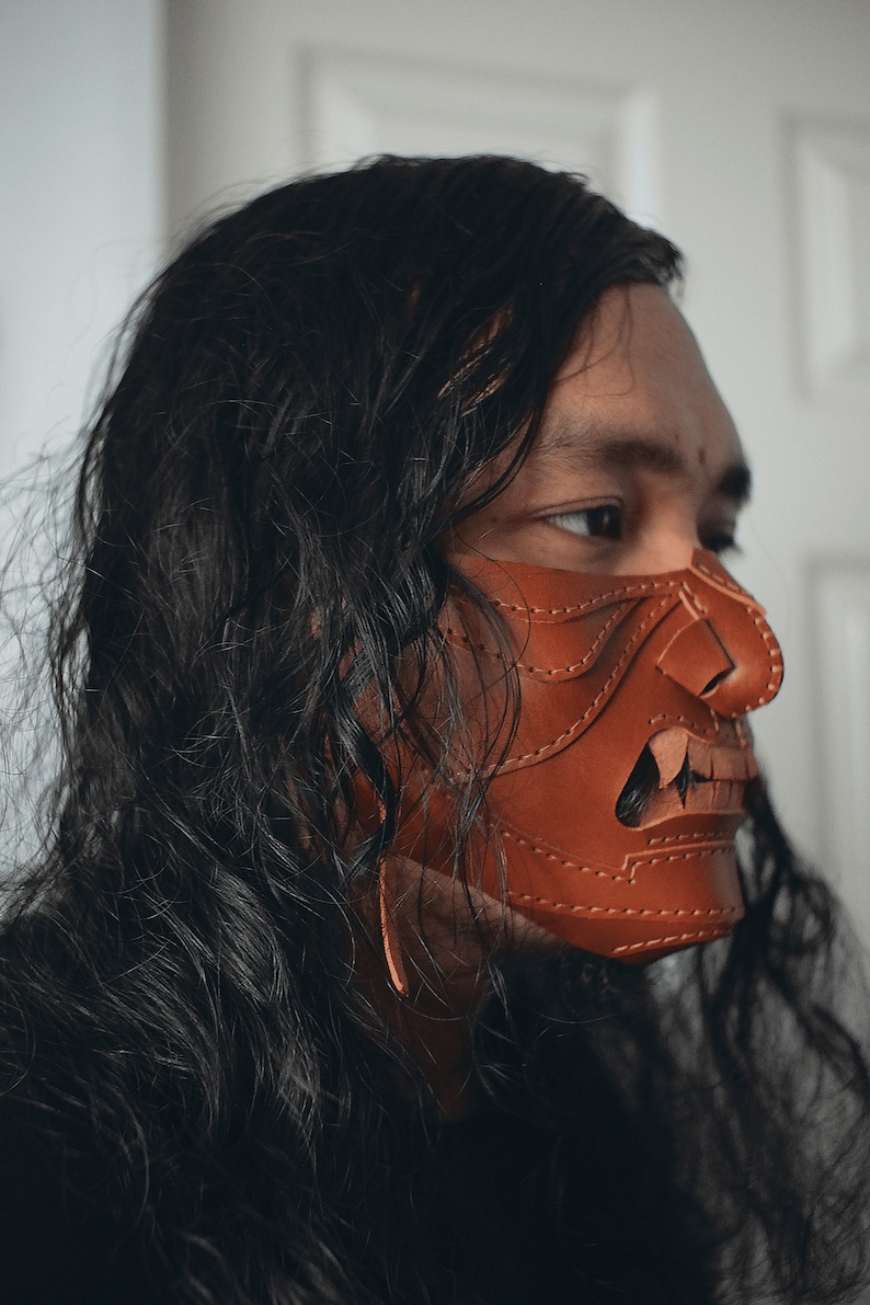 Pu&ograve; includere: Una persona con lunghi capelli neri indossa una maschera di cuoio marrone con cuciture intricate e un ghigno pieno di denti.