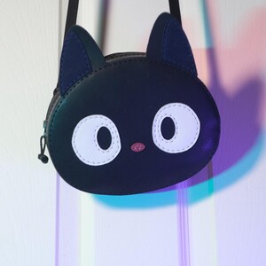 Puede incluir: Bolso bandolera negro con forma de cara de gato, con ojos blancos grandes y nariz rosa. El bolso tiene una correa y orejas negras, y un cierre de cremallera. El bolso est&aacute; colgado de una puerta blanca.