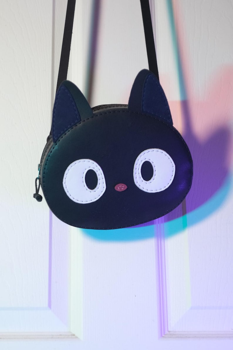 Puede incluir: Bolso bandolera negro con forma de cara de gato, con ojos blancos grandes y nariz rosa. El bolso tiene una correa y orejas negras, y un cierre de cremallera. El bolso est&aacute; colgado de una puerta blanca.