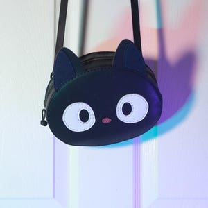 Puede incluir: Bolso bandolera negro con forma de cara de gato, con grandes ojos blancos, una nariz rosa y orejas azul oscuro. El bolso tiene una correa negra y un cierre de cremallera. El dise&ntilde;o es caprichoso y adecuado para objetos peque&ntilde;os.