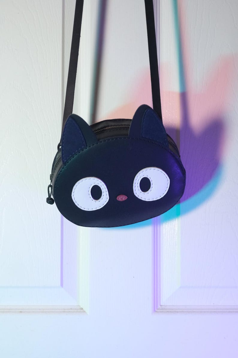 Puede incluir: Bolso bandolera negro con forma de cara de gato, con grandes ojos blancos, una nariz rosa y orejas azul oscuro. El bolso tiene una correa negra y un cierre de cremallera. El dise&ntilde;o es caprichoso y adecuado para objetos peque&ntilde;os.