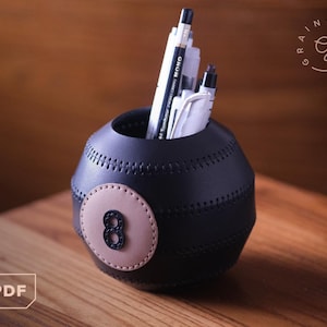 May include: A black leather pen holder shaped like an eight ball, with a tan circle and the number 8. The holder is filled with pens and pencils. The item is on a wooden surface.