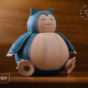 Puede incluir: Un peluche de cuero azul y beige hecho a mano, que se asemeja a un personaje con los ojos cerrados y orejas pequeñas. El juguete está cosido con hilo visible y tiene pies pequeños. El logotipo PDF está en la esquina inferior izquierda.