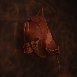 Pu&ograve; includere: Una maschera in pelle marrone con cuciture intricate e un design ritagliato. La maschera &egrave; appesa a un cinturino in pelle.