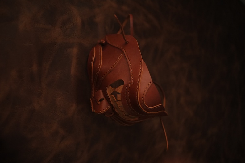 Pu&ograve; includere: Una maschera in pelle marrone con cuciture intricate e un design ritagliato. La maschera &egrave; appesa a un cinturino in pelle.