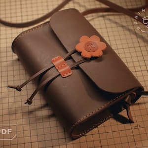 Könnte beinhalten: Eine braune Leder-Umhängetasche mit Klappenverschluss und einem dekorativen Blumenakzent. Die Tasche hat einen Riemen und ein kleines Fach auf der Vorderseite. Die Tasche befindet sich auf einer karierten Oberfläche.
