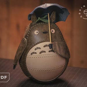 May include: A handmade leather Totoro figure with a blue umbrella. The figure is mainly brown and cream, with stitched details and a small green leaf. The design is inspired by the animated film.