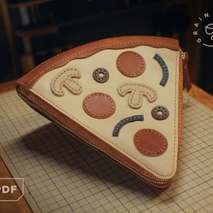 Puede incluir: Una bolsa de cuero marrón con cremallera en forma de rebanada de pizza con fondo blanco y diversas aplicaciones de cuero de colores. La bolsa tiene una cremallera y el texto "GRAINY DAYS" está en el lateral.