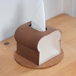 Pode incluir: Uma caixa de len&ccedil;os de papel castanha e branca em forma de fatia de p&atilde;o, com um len&ccedil;o branco a sair do topo. A caixa de len&ccedil;os est&aacute; assente numa base redonda de corti&ccedil;a, colocada numa superf&iacute;cie de madeira.