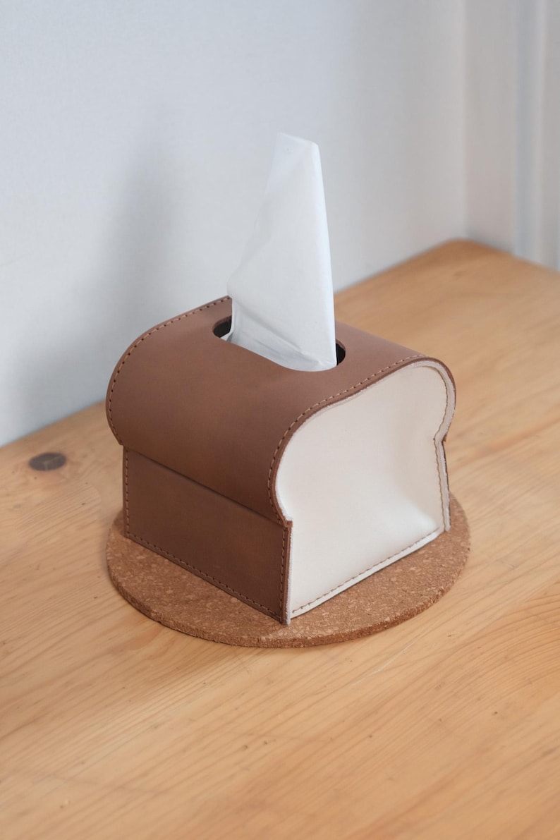 Pode incluir: Uma caixa de len&ccedil;os de papel castanha e branca em forma de fatia de p&atilde;o, com um len&ccedil;o branco a sair do topo. A caixa de len&ccedil;os est&aacute; assente numa base redonda de corti&ccedil;a, colocada numa superf&iacute;cie de madeira.