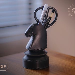 Puede incluir: Un organizador de escritorio de cuero negro con forma de caballo de ajedrez, que contiene tijeras, un bolígrafo blanco y un bolígrafo negro. El organizador tiene una base escalonada y un diseño cosido. El fondo está borroso, con una ventana y una superficie de madera.