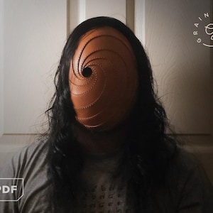 May include: A brown leather mask with a spiral design and a central eye hole. The mask is worn by a person with long black hair and a gray t-shirt. The mask is the focal point of the image.