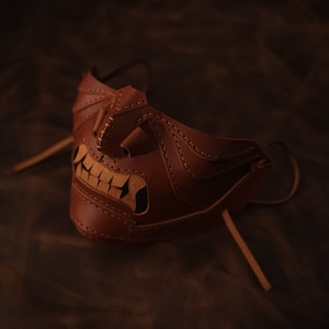 Pu&ograve; includere: Una maschera in pelle marrone con un design cucito che ricorda un viso con un ghigno pieno di denti. La maschera ha un lungo laccio in pelle per legarla intorno alla testa.