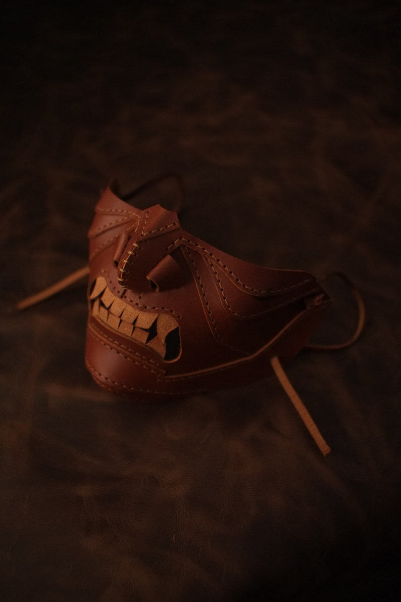 Pu&ograve; includere: Una maschera in pelle marrone con un design cucito che ricorda un viso con un ghigno pieno di denti. La maschera ha un lungo laccio in pelle per legarla intorno alla testa.