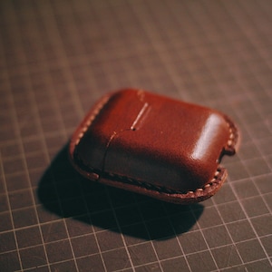 May include: A brown leather case for AirPods or other small electronics. The case has a stitched seam around the edge and a flap that closes with a snap.