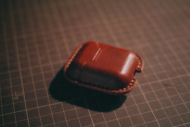 May include: A brown leather case for AirPods or other small electronics. The case has a stitched seam around the edge and a flap that closes with a snap.