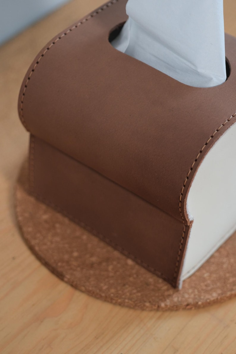 Pode incluir: Uma caixa de len&ccedil;os de couro marrom e branco. A se&ccedil;&atilde;o superior &eacute; marrom, com um design arredondado e uma fenda para dispensar len&ccedil;os. A se&ccedil;&atilde;o inferior &eacute; branca. A caixa est&aacute; em uma base de corti&ccedil;a.