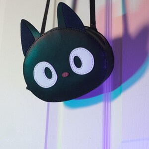 Puede incluir: Bolso bandolera negro con forma de gato con un compartimento redondo con cremallera. El bolso presenta ojos ovalados blancos, una nariz rosa y orejas negras. El bolso cuelga de una correa negra.