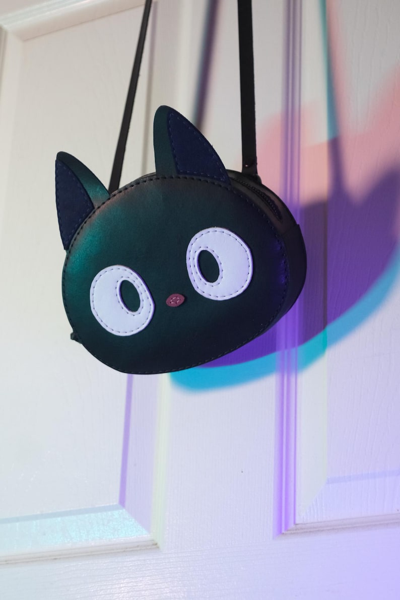 Puede incluir: Bolso bandolera negro con forma de gato con un compartimento redondo con cremallera. El bolso presenta ojos ovalados blancos, una nariz rosa y orejas negras. El bolso cuelga de una correa negra.