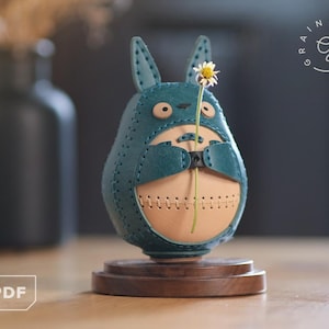 May include: A teal leather Totoro-shaped vase holding a small white and yellow flower. The vase is stitched together with tan thread and sits on a wooden base. The vase has a tan interior and is on a wooden base.