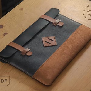 Puede incluir: Funda de tableta de cuero negro y marrón con correas de cuero marrón. La funda tiene un panel de aspecto de gamuza y un pequeño parche cuadrado con dos ranuras paralelas. El logotipo "Rainy Days" está en la esquina superior derecha.