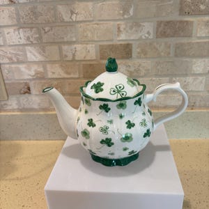 May include: White ceramic teapot with a green shamrock pattern and accents. The teapot has a rounded body, a curved spout, and a handle. The lid is topped with a green knob. The teapot is sitting on a white surface.