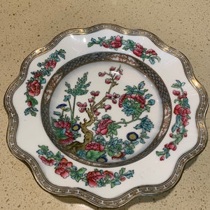 May include: A white porcelain bowl with a scalloped edge and a gold trim. The bowl features a floral design with pink, blue, and green flowers and leaves. The design is centered around a tree with a brown trunk and branches.