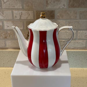 May include: A white and red striped teapot with a gold-colored lid and handle accents. The teapot is placed on a white surface, showcasing its elegant design. The teapot's design features vertical red stripes alternating with white stripes.