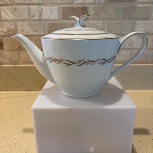 May include: White porcelain teapot with a lid, gold trim, and a decorative floral pattern. The teapot has a curved spout and handle. The lid features a decorative finial. The teapot is sitting on a white block.