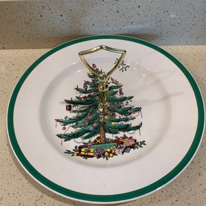May include: A white ceramic serving plate with a green border and a gold handle. The plate features a Christmas tree design with ornaments and presents.