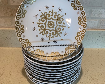 May include: A stack of 12 white ceramic dinner plates with a gold and white geometric pattern. The plates are stacked on a white countertop.