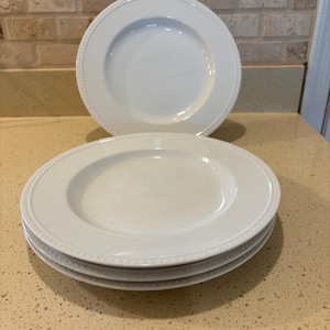 May include: A stack of four white ceramic plates with a beaded rim design. One plate is stacked on top of the others. Another plate is placed behind the stack. The plates are on a light-colored countertop.